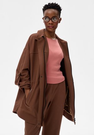 Kobieta w okrągłych okularach, różowym topie oraz oversize'owej brązowej kurtce i spodniach, stojąca z rękami w kieszeniach, patrząca na bok.