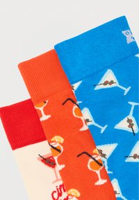 Τρεις πολύχρωμες κάλτσες με σχέδια ποτηριών κοκτέιλ σε κόκκινους, πορτοκαλί και μπλε τόνους, με ποτά που έχουν καλαμάκια, ελιές και φέτες λεμονιού.