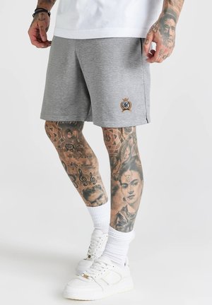 Hombre con pantalones cortos grises con logo bordado, calcetas blancas, zapatillas blancas y tatuajes detallados en las manos y piernas.