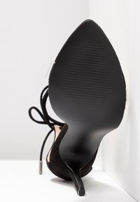 Chaussure noire à talon haut pointu avec semelle texturée et bride de cheville nouée en nœud, vue par le dessous sur fond blanc.