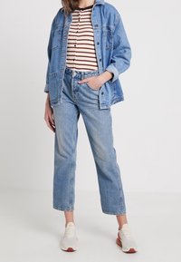 Hellblaue Jeansjacke über einem gestreiften, geknöpften Oberteil, kombiniert mit hoch taillierten hellblauen Jeans und weißen Turnschuhen mit roten Akzenten.