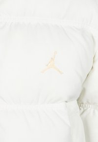 White puffy jacket with elastic cinching at the waist. Features a stitched gold Jumpman logo on the chest. Smooth texture.