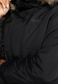 Personne portant une veste isolante Didrikson noire avec bordure en fausse fourrure et cheveux tressés, main reposant près du visage.