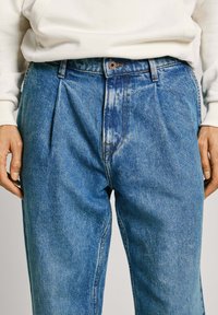 Ljusblå denimbyxor med veck fram, knappstängning och sidofickor. Tyget har en lätt blekning och struktur.