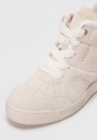 Beige Wildleder High-Top Sneaker mit weißen Schnürsenkeln, perforierter Zehenpartie und goldenen Logodetails. Verfügt über eine hellfarbige Gummisohle.