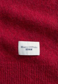 Rode gebreide stof met een zachte textuur. Een wit label met de tekst "Marc O'Polo DENIM" in donkerblauwe letters, prominent op het oppervlak geplaatst.