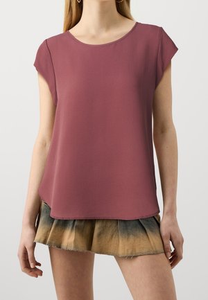 Vrouw die een kortemouws stoffig roze top en een geplooide minirok met een bruin-zwart verloopmotief draagt.