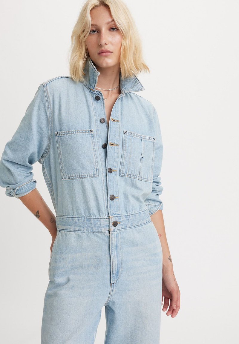 Lyseblå denim-dragt med høj krave, to frontlommer, knaplukning og optrukne ærmer; har en afslappet pasform og falmet tekstur.