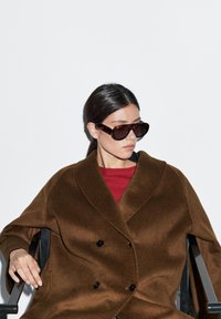 Brązowy wełniany płaszcz w podwójnie zapinanym fasonie, z czarnymi guzikami, noszony na czerwonym swetrze. Duże tortoise'owe okulary przeciwsłoneczne dopełniają stylizację.