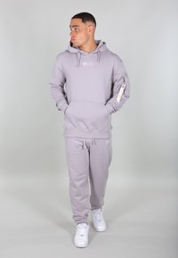 Svetlo šedá mikina s kapucňou a vreckom na prednej strane, doplnená o zipsové vrecko na rukáve, v kombinácii s ladnými teplákmi a bielymi teniskami.