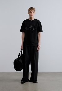 Μαύρο μπλουζάκι με το λογότυπο "Hugo", μαύρο παντελόνι σε χαλαρή γραμμή, κρατώντας μια μαύρη στρογγυλή τσάντα. Μινιμαλιστικός σχεδιασμός, απαλό ύφασμα, χωρίς ορατά σχέδια.