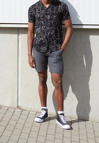 Man met een zwart shirt met witte abstracte gezichten, grijze shorts, witte sokken en zwarte hoge sneakers staat tegen een betonwand.