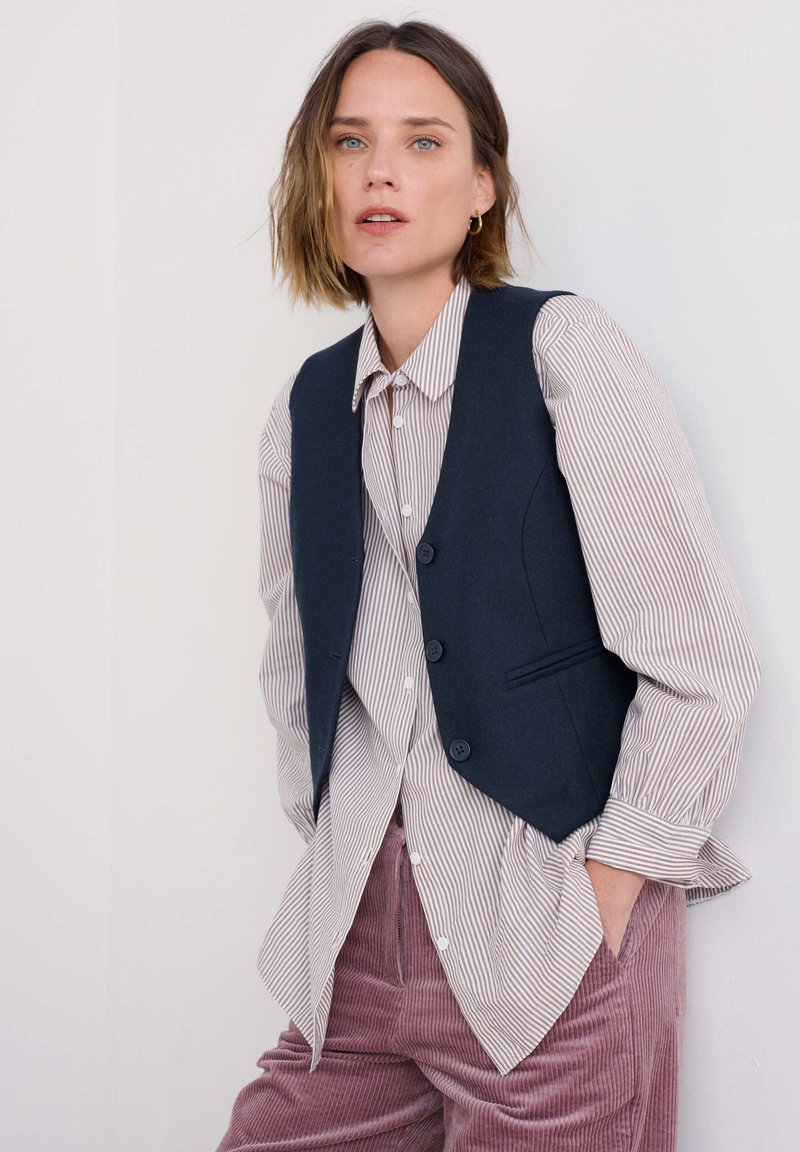 Tmavomodrý vest s tromi gombíkmi a bočnými vreckami, vrstvený cez prúžkovanú košeľu, skombinovaný s ružovými kordovým nohavicami. Celkový hladký vzhľad.
