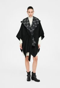 Poncho nero con colletto patterned, dotato di una cintura a fiocco. Il colletto interno presenta un design progettato, abbinato a stivali neri alla caviglia.