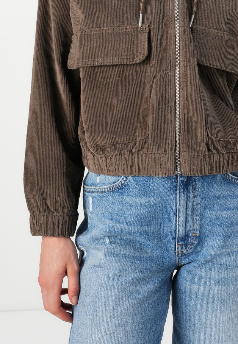 Veste en velours côtelé marron avec fermeture éclair, dotée de deux poches poitrine et d'un bas élastiqué. Portée sur un jean en denim délavé bleu clair.