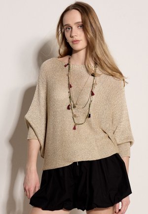 Junge Frau mit langen Haaren, die einen beigen Strickpullover, eine mehrschichtige Perlenkette und einen lockeren schwarzen Rock trägt, steht vor einem schlichten Hintergrund.