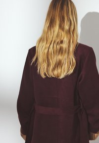 Burgundia sametist mantel, millel on vöökoht, laiad varrukad ja lihtne disain. Pikk, laineline blonde juuksed on nähtavad.