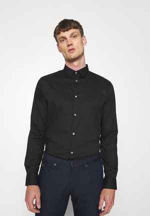 Chemise noire à manches longues avec un col boutonné, présentant un logo discret, des boutons brillants et une texture de tissu lisse. Associée à un pantalon sombre.