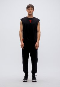 Μαύρο αμάνικο πουκάμισο με κόκκινο γραφικό στο στήθος, σε συνδυασμό με μαύρο κωνικό φούτερ και μαύρα αθλητικά παπούτσια. Απλός, αθλητικός σχεδιασμός.