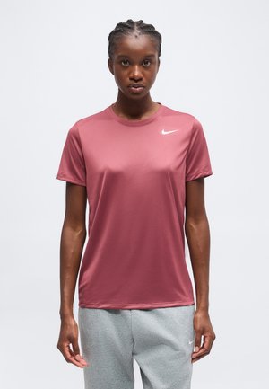 Mladá žena so zapletenými vlasmi, oblečená v ružové ružové tričko Nike s krátkym rukávom a svetlosivých teplákoch, tváriaca sa dopredu na bielom pozadí.