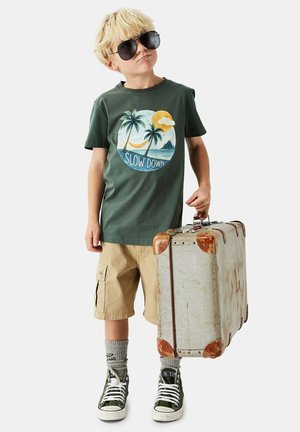 Lyshåret dreng iført solbriller, grøn t-shirt med teksten "Slow Down", beige shorts og sneakers, der holder en vintage kuffert med en neutral baggrund.