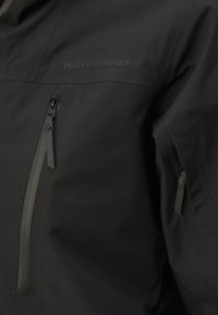 Veste noire imperméable avec une texture lisse, dotée d'un col montant, de poches zippées et d'un logo brodé discret sur la poitrine.