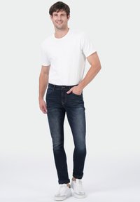 Homme debout avec une main dans la poche, portant un t-shirt à manches courtes blanc, un jean bleu foncé et des baskets blanches sur un fond uni.