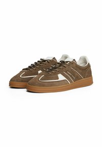 Sneaker low-top in suede marrone con un accento in pelle bianca, punta tonda, suola in gomma testurizzata e lacci scuri. Logo visibile di lato.