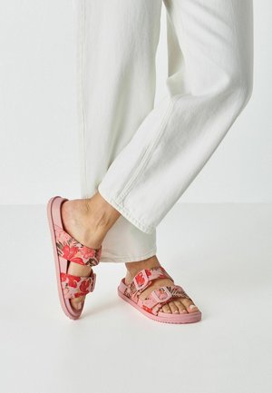 Pieds portant des sandales à double bride à fleurs roses associées à un pantalon blanc, debout sur un sol clair.