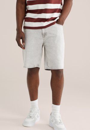 Mand iført lysegrå denimshorts, hvide sneakers, hvide sokker og en vinrød og hvidstribet skjorte, stående mod en neutral baggrund.