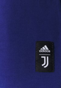 Un tejido azul con una textura suave presenta un parche de logo negro con texto blanco: "adidas" sobre "JJ" en el lado derecho.