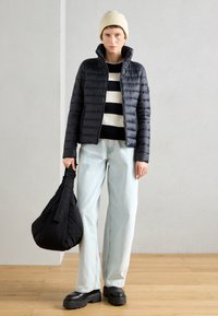 Melna pūkaina jaka ar augstu apkakli, svītrainā džemperī, gaiši zilos džinsos un masīvās melnās platformas kurpēs, tur melnu somu.