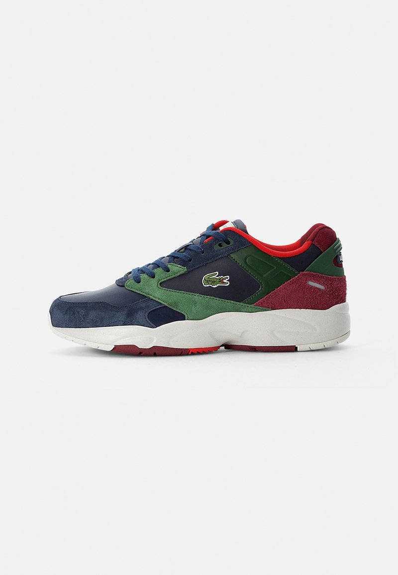Baskets basses avec panneaux en daim bleu marine, vert et bordeaux, doublure intérieure rouge, semelle blanche et logo crocodile Lacoste sur le côté.