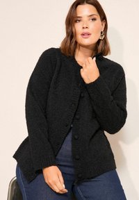 Frau mit schulterlangem braunem Haar, die goldene Creolen, eine schwarze, flauschige, geknöpfte Strickjacke und blaue Jeans trägt, posiert vor einem neutralen Hintergrund.