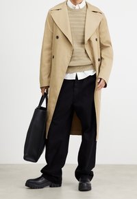 Beige Doppelreiher-Trenchcoat, gerippter beiger Pullover, weißes Hemd mit Kragen, schwarze weite Hose und klobige schwarze Gummistiefel.