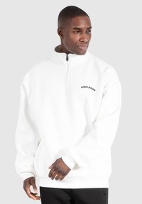 Pullover bianco in pile con colletto a zip e polsini a costine, caratterizzato da un logo nero sul petto. Texture morbida e vestibilità comoda.