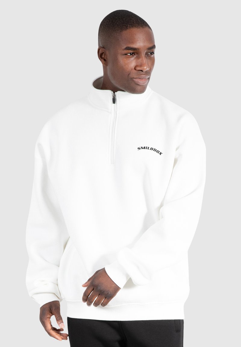 Pullover bianco in pile con colletto a zip e polsini a costine, caratterizzato da un logo nero sul petto. Texture morbida e vestibilità comoda.