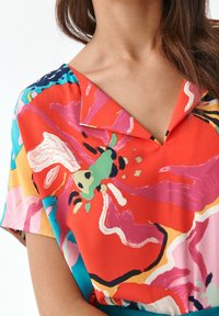 Kolorowa koszula w kwiaty z dekoltem w serek, z żywym wzorem w odcieniach czerwieni, różu, zieleni i niebieskiego. Wykonana z lekkiego materiału, krótkie rękawy.