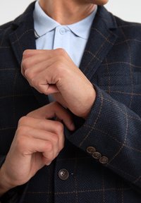 Jūros jėgos herringbone estilo švarkas su oranžiniu patikrinimo raštu, trys rudi mygtukai ant rankovės, derinama su šviesiai mėlynu marškinėliais su apykakle.