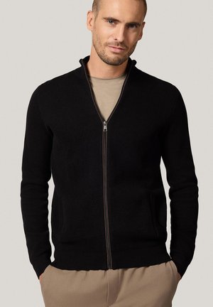 Mężczyzna w czarnej, strukturalnej bluzie na zamek, noszący beżową koszulę oraz z rękami w kieszeniach beżowych spodni, na tle jednolitym.