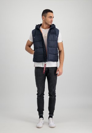 Marineblauer Steppweste mit Kapuze und seitlichen Taschen, ausgestattet mit einem Reißverschluss und kontrastierender Innenauskleidung, getragen über einem weißen T-Shirt und schwarzen Jeans.