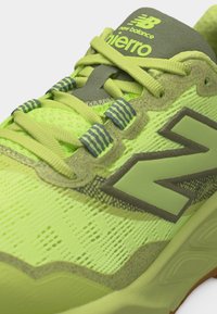 Κοντινή λήψη νεον πράσινου παπουτσιού τρεξίματος New Balance με χακί λεπτομέρειες, λεπτομερή κορδόνια και λογότυπο "Fierro" στη γλώσσα.