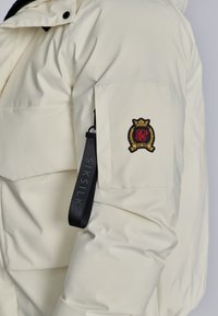 Giubbotto piumino color crema con texture trapuntata, dotato di un logo a patch sulla manica sinistra e di una striscia nera con il marchio sulla tasca.