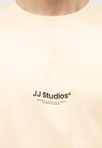 Μπεζ μπλουζάκι με μαύρο κείμενο "JJ Studios" και μικρότερο κείμενο "Παρασχεθεί από την ομάδα J&J, Σχεδιασμένο στη Δανία" στο στήθος.