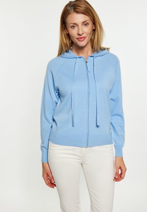 Hellblaues Zip-Hoodie aus weichem Strickstoff, mit Kapuze, Kordelzug und gerippten Bündchen, kombiniert mit weißen Hosen.