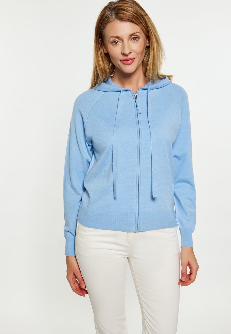Felpa leggera azzurra con zip in un morbido tessuto a maglia, con cappuccio, coulisse e polsini a coste, abbinata a pantaloni bianchi.