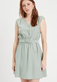 Robe verte sans manches avec un décolleté en V, fermeture à boutons et taille ceinturée. Fabriquée en tissu lisse avec une jupe fluide.