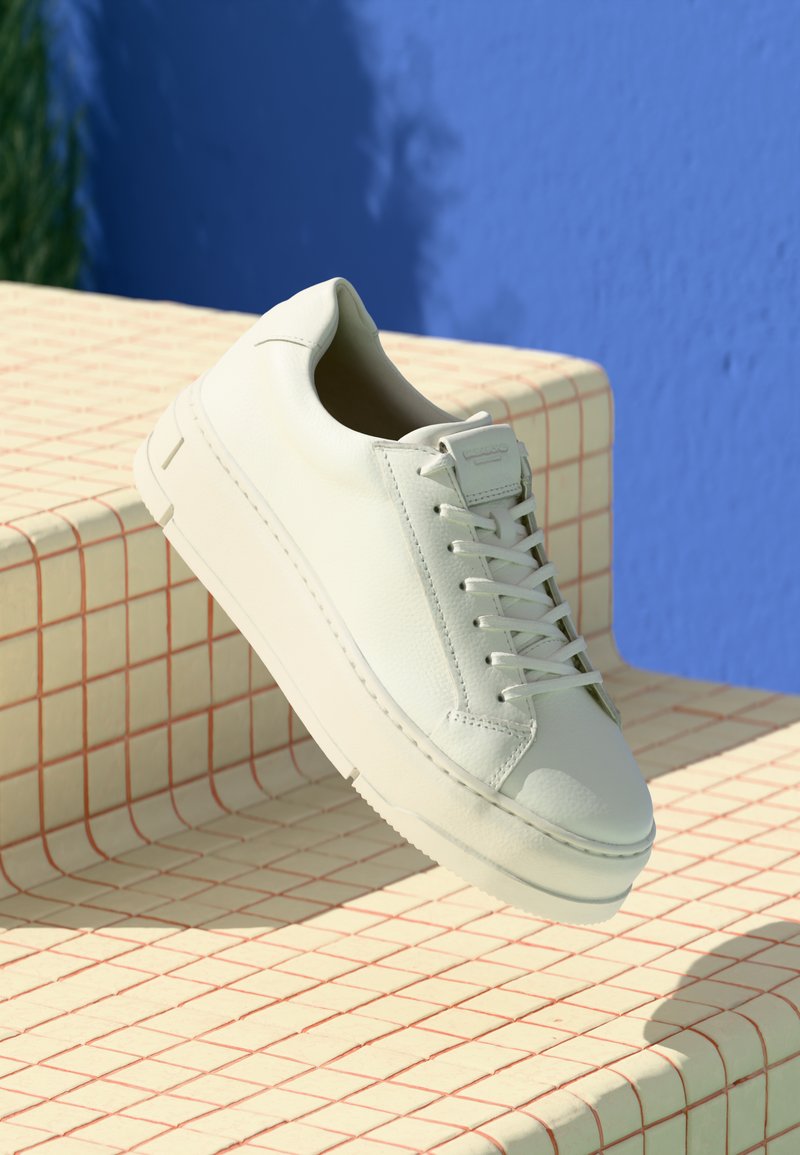 White leather sneaker with round toe, textured upper, and flat laces. Features a thick rubber sole and minimalistic design against a patterned backdrop.