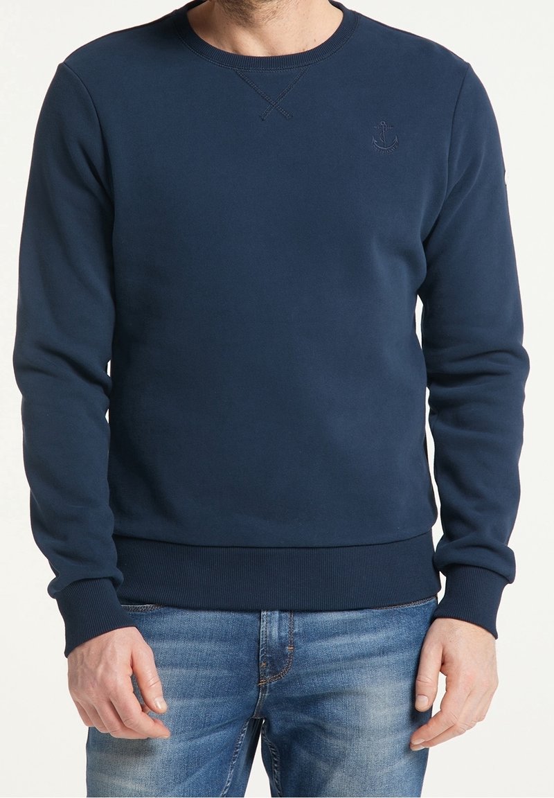 Homme portant un sweat-shirt bleu marine à col rond avec un logo d'ancre sur la poitrine et un jean bleu, debout devant un fond blanc.