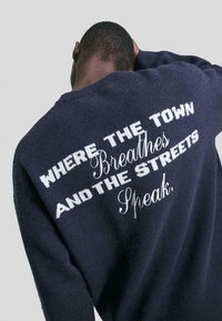 Oseba v mornarsko modrem puloverju s belim besedilom na hrbtni strani: "KJER MESTO DIHA IN ULICE GOVORIJO."